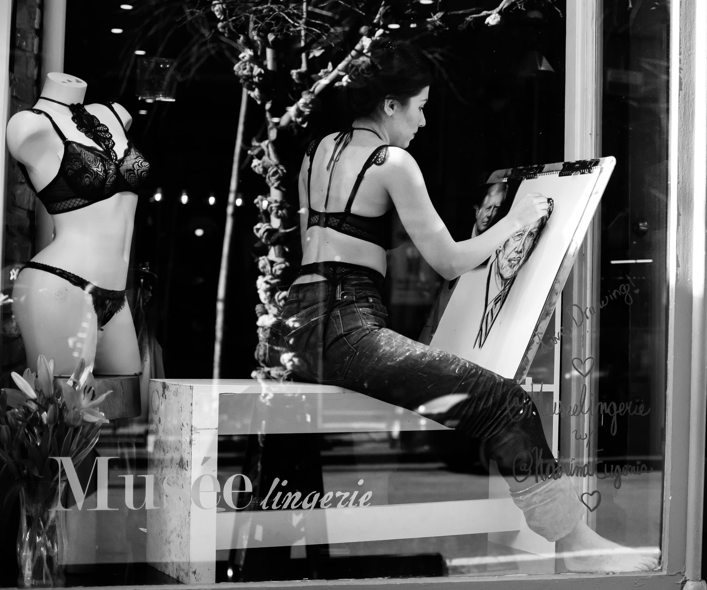 A woman sits inside a lingerie boutique by the window, drawing on an easel while studying a reference picture, black and white