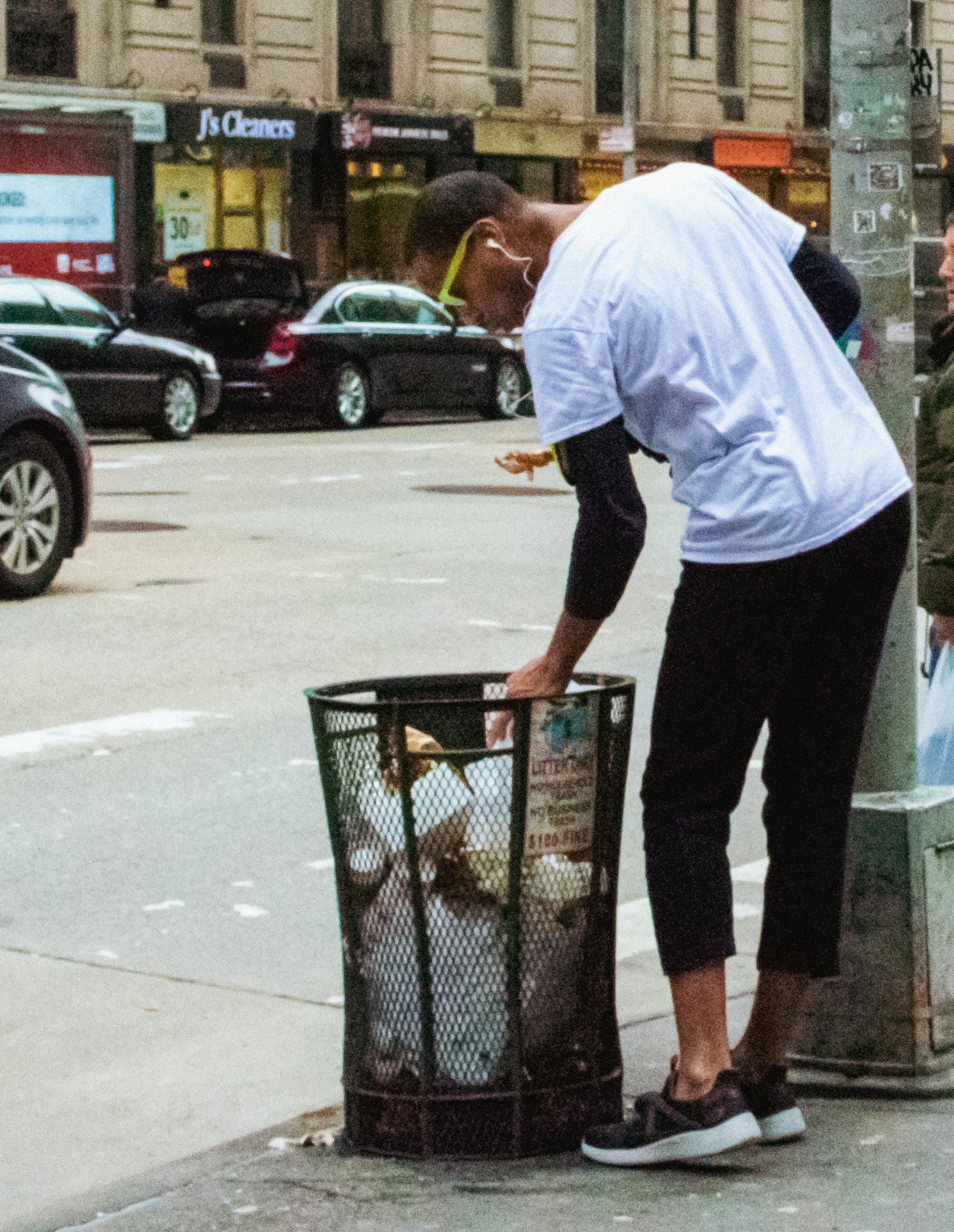 A person in a white t-shirt and yellow sunglasses searching through a mesh street trash can on a New York City sidewalk