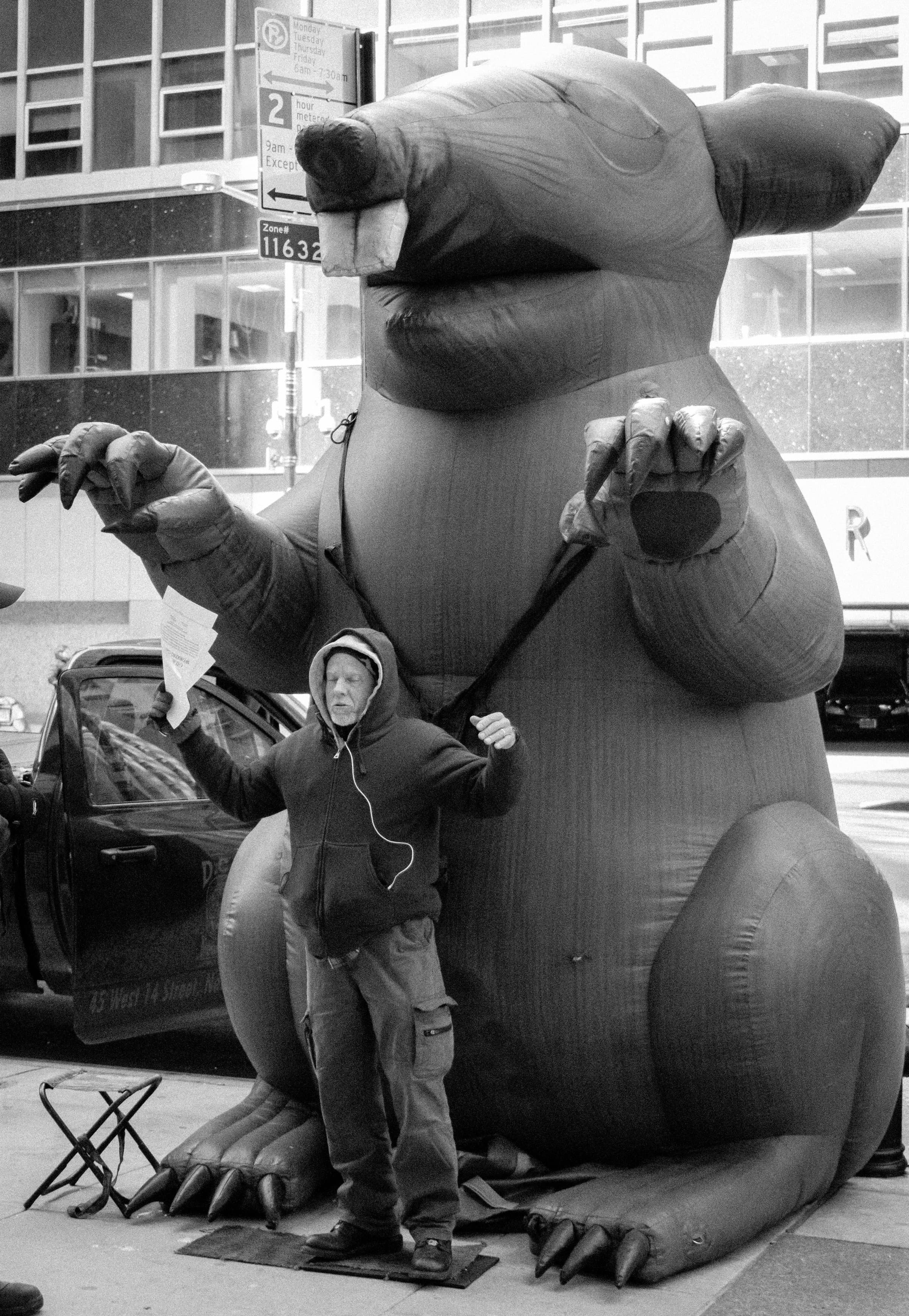 A man in a hoodie holds the tether of a giant inflatable union protest rat on a Manhattan sidewalk.