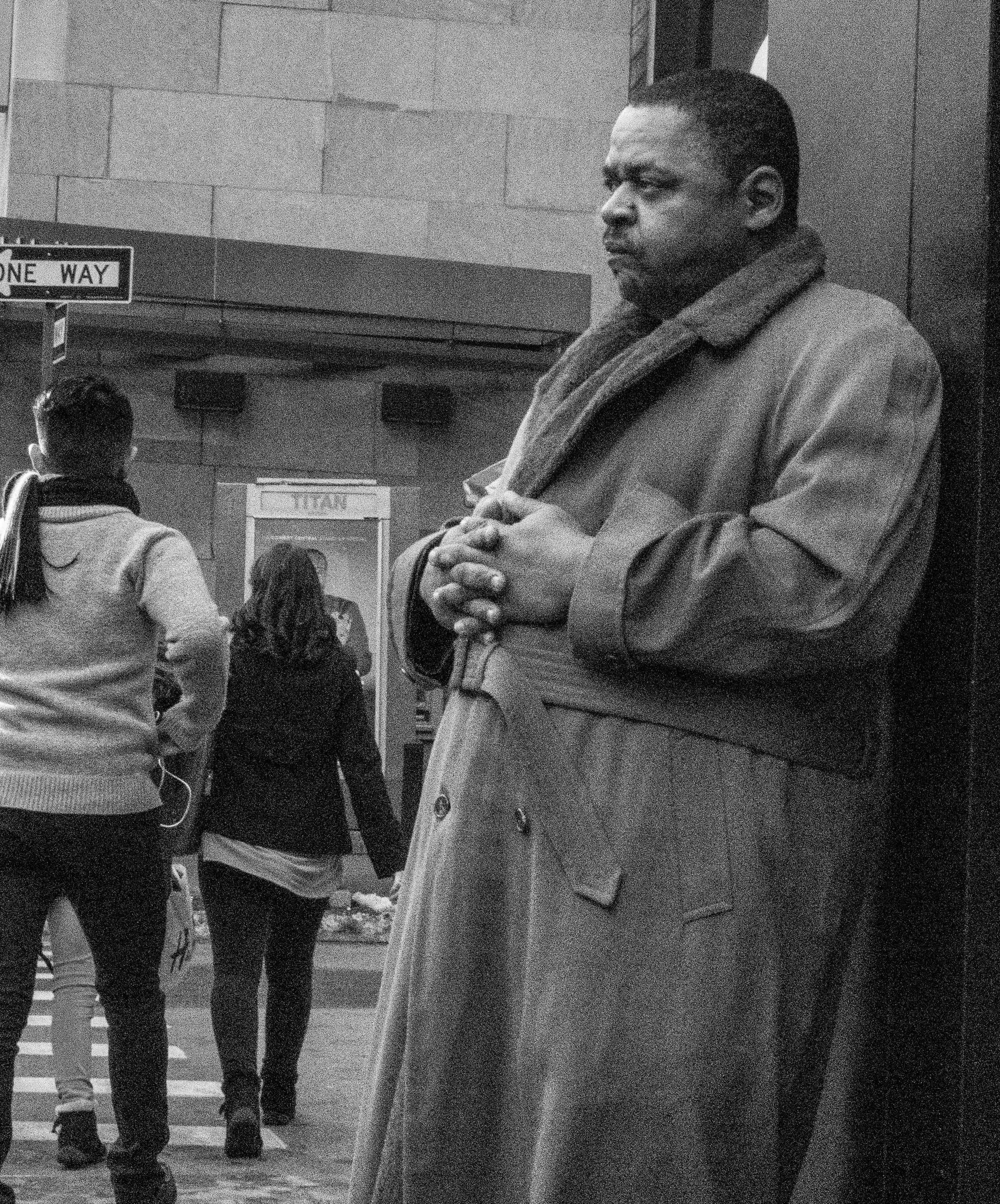A man stands still against a wall on a busy New York sidewalk, hands clasped, watching
