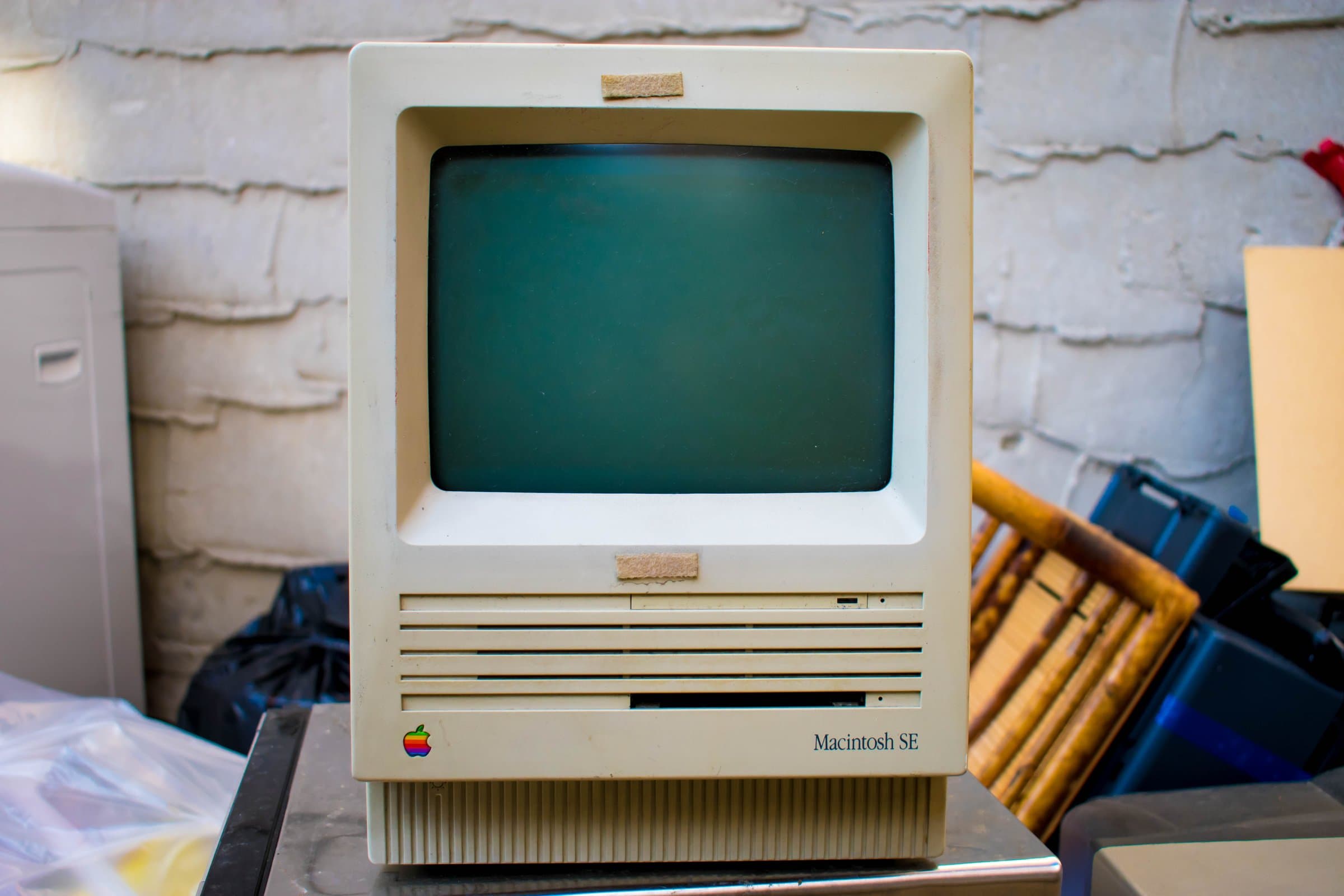 A Macintosh SE sits dormant among junk and trash bags against a white brick wall.
