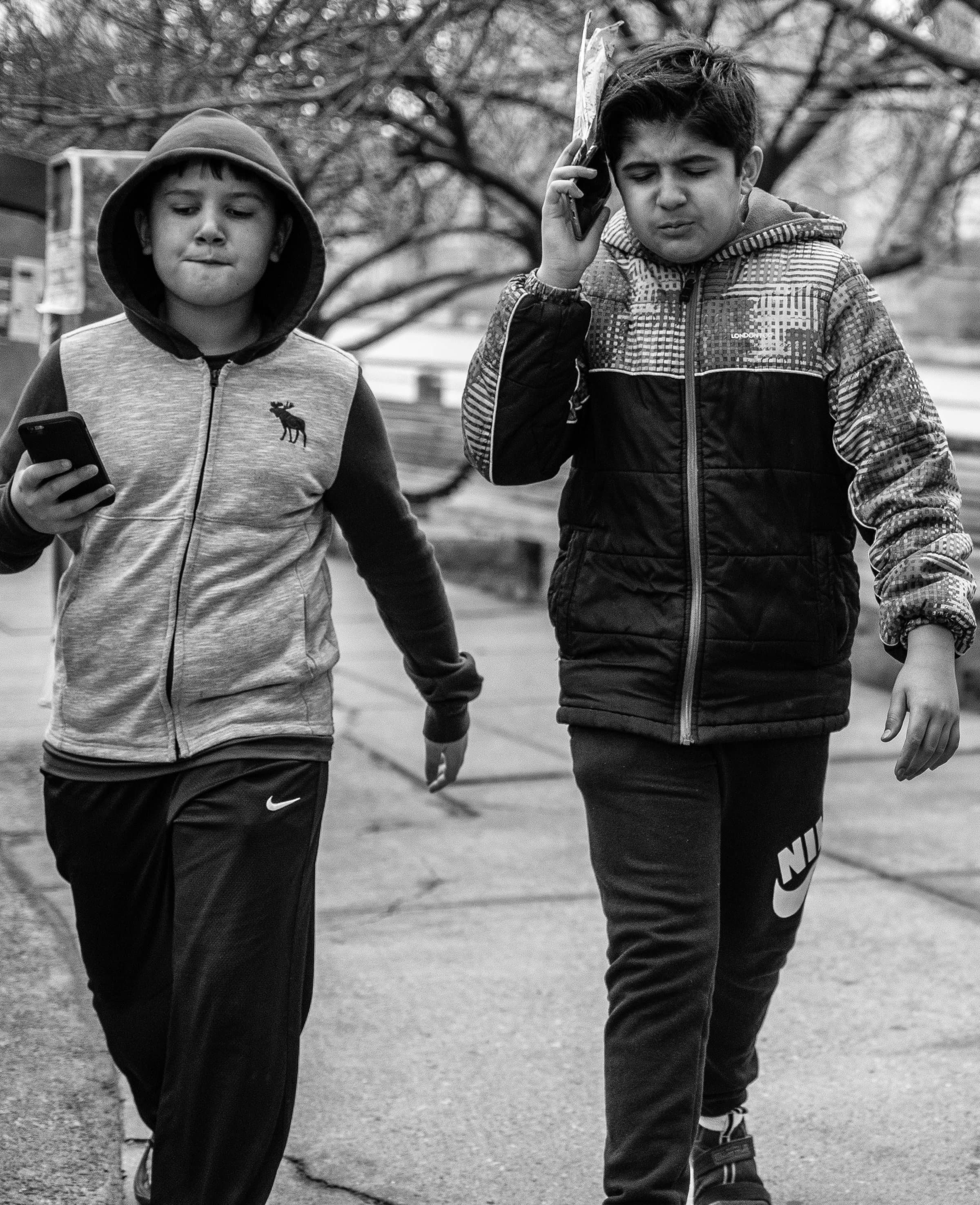 Two kids in hoodies walk with phones in hand on a path through bare winter trees.
