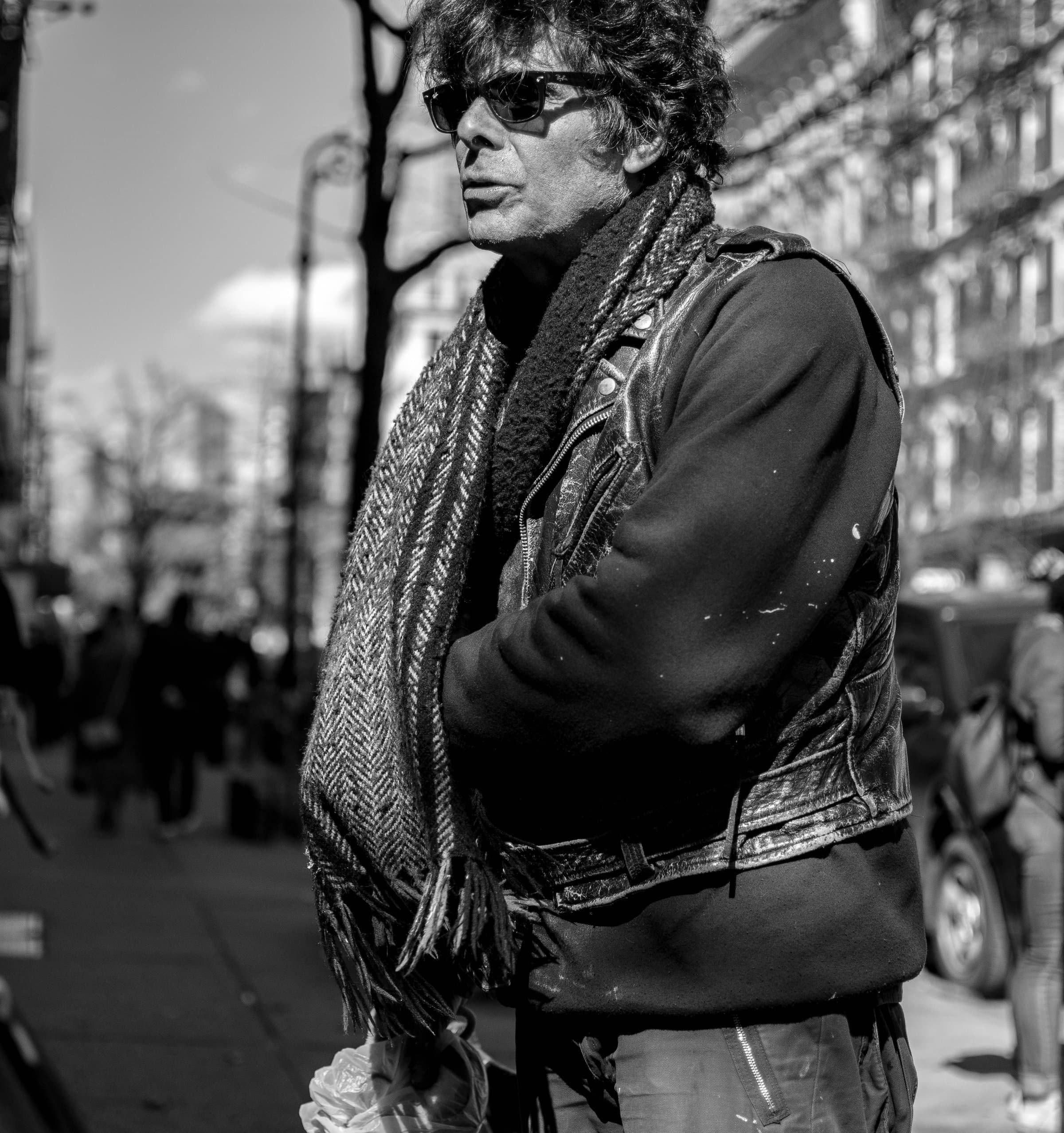 An older man in sunglasses, leather jacket, and scarf surveys a crowded city street, his gaze direction hidden behind dark lenses, black and white