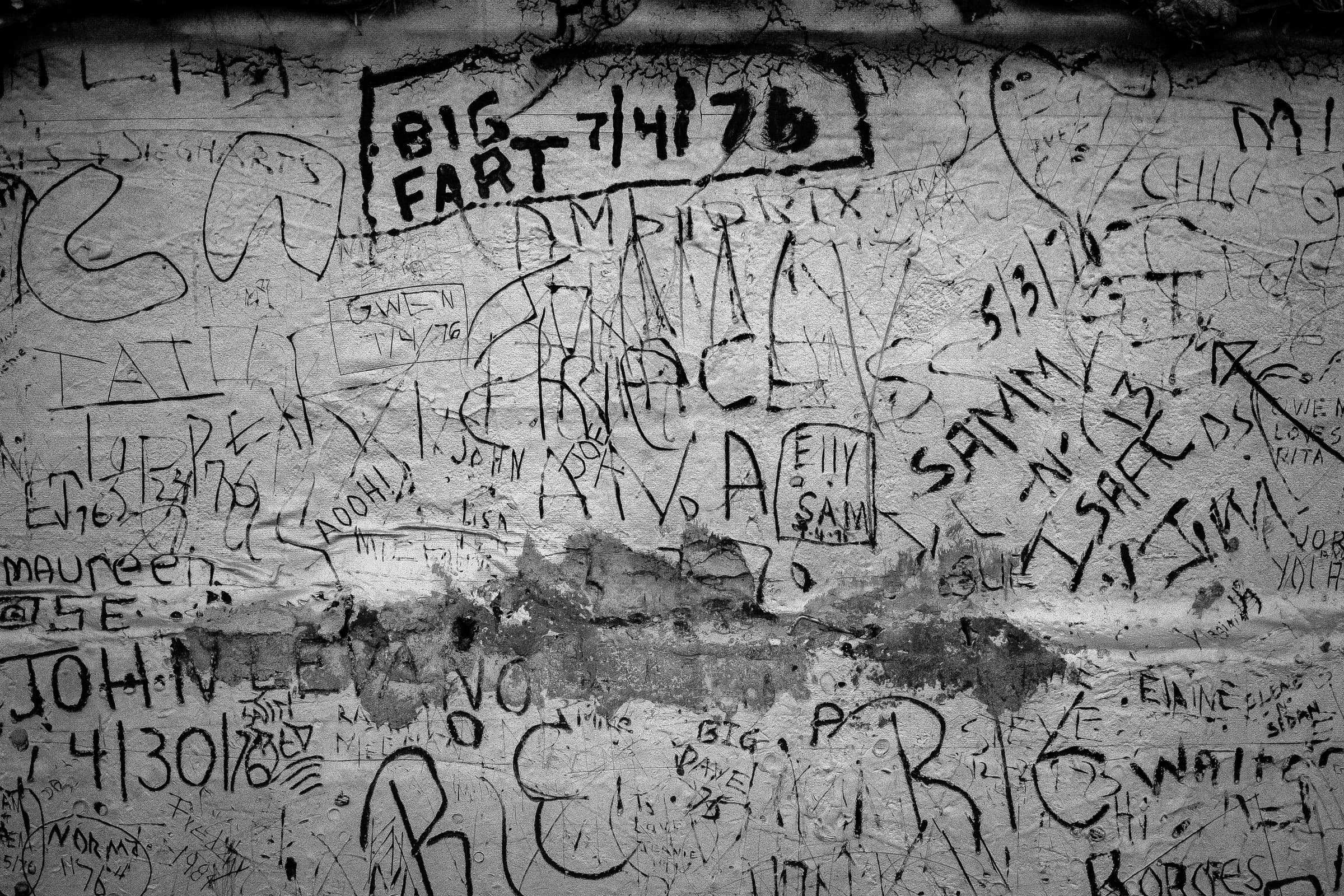A New York wall densely covered in scratched names and handwritten dates spanning decades, black and white