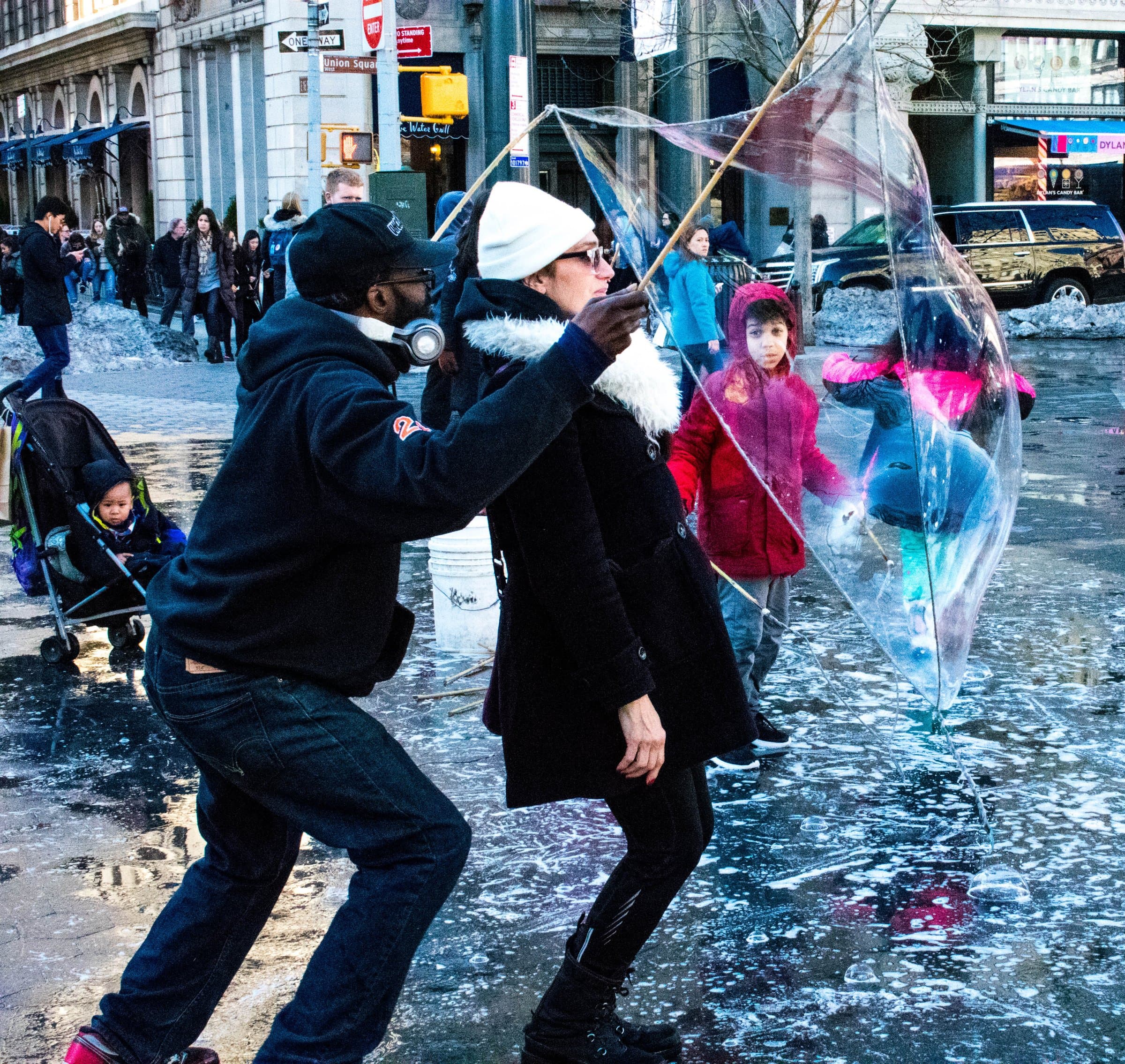 Two people maintain a giant soap bubble on a Manhattan sidewalk while a crowd watches in winter